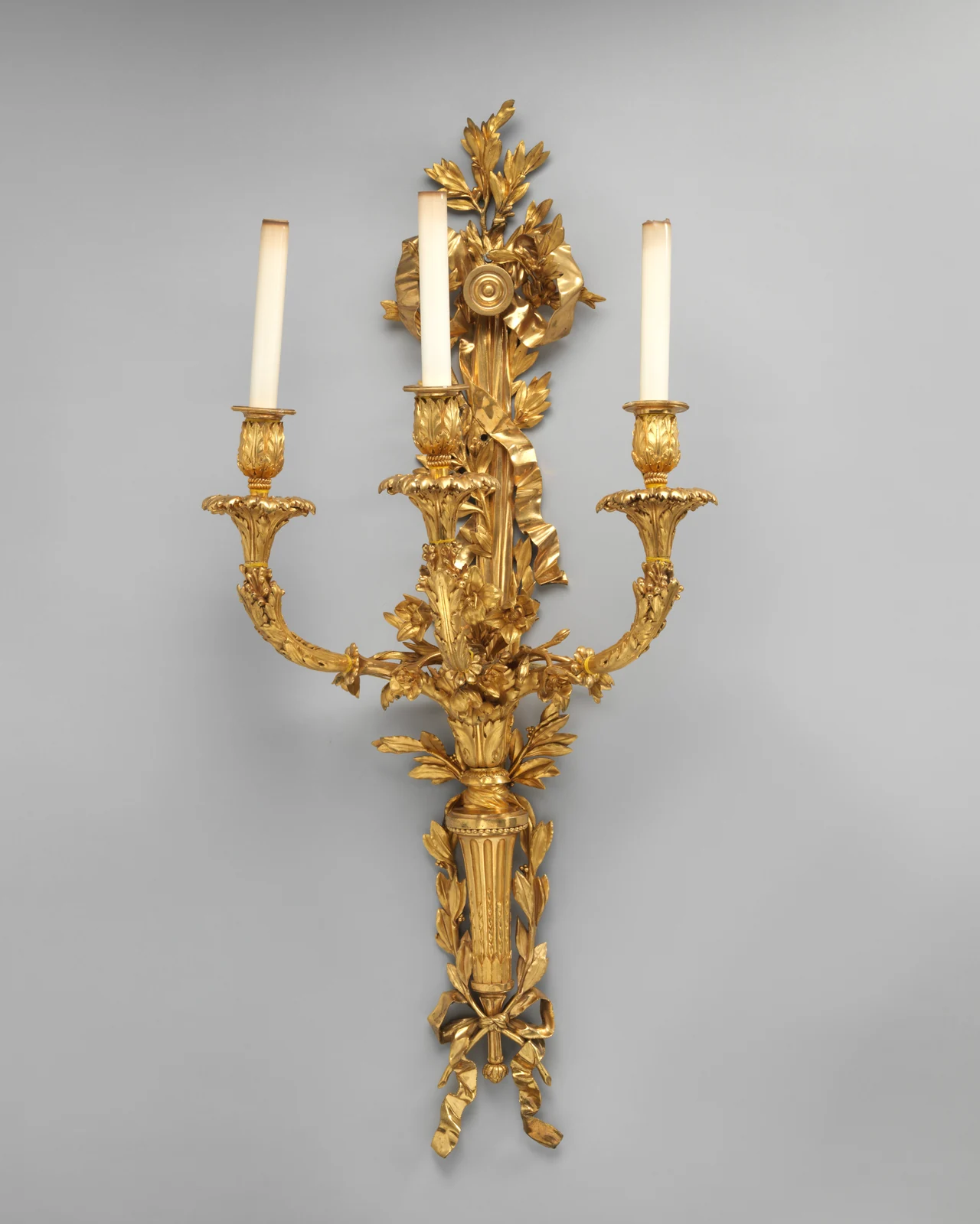 Aplique parisino de tres brazos de hacia 1785, con antorcha acanalada, lazos y hojas de laurel en bronce dorado